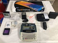 Photo showing all items: Sega Dreamcast Keyboard box, Brother PT-320 labelling system with power source and manual, Power Mat charger with carrying case and adapter, Nomis battery pack, digital calendar and envelope opener with manual.