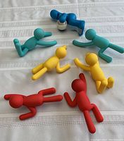 Overview of six colorful Umbra Buddy hooks laid out on fabric. Includes teal, blue, yellow, and red humanoid figure hooks, and one blue tape dispenser shaped like legs.