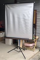 Full view of the white projection screen set up on the tripod stand in an indoor storage space with some surrounding boxes and furniture.