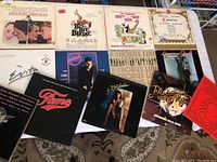 Top-down view of 11 vintage vinyl LP covers laid on carpet. Visible titles include Evita, Fame, Flashdance, A Chorus Line, American Gigolo, Camelot, Doctor Zhivago, Cocktail, FM, Doctor Dolittle with Rex Harrison, and Bye Bye Birdie.