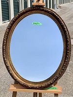 Full front view of the oval mirror showing the entire reflective surface and ornate gold-tone frame with a floral and scroll pattern design.