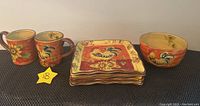 Photo showing two large mugs with rooster and sunflower motifs alongside four square decorative plates with rooster and floral designs.