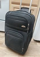 Black Samsonite upright suitcase standing on the floor, showing front compartments and handle extended.