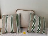 Three outdoor pillows arranged on a surface, showing one solid beige pillow and two striped pillows with green and grey vertical stripes.