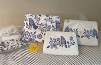 Three white fabric zippered pouches of different sizes with blue bird and floral embroidery, one with a gold 'P' emblem patch, displayed on a black and white patterned surface.