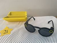 Front view of black Snapchat Spectacles with yellow charging case in background on white perforated cloth surface.