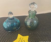 Two vintage glass perfume bottles placed on black fabric; one aqua blue with rounded stopper and ribbed body, and one green with faceted stopper and etched design.