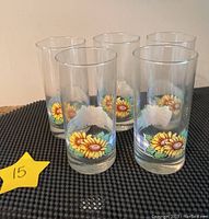 Photo showing five tall clear glasses with sunflower motifs and blue checkered base pattern in an upright position on black textured surface.