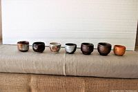 Seven small Japanese ceramic cups, arranged in a line, showing a range of glazes and styles, placed on a cloth surface with a white backing.