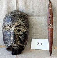 Front view of hand carved wooden Timor mask with attached facial hair and accompanying carved lanceolate wooden implement with two notches and decorative incisions.
