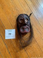 Photo of a dark brown wooden First Nations mask with carved facial features lying on a wooden floor. The mask has dark hair attached around the sides and chin area. A lot number card is placed beside it for scale.