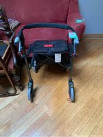 Front view of the foldable walker showing padded seat, hand grips, and wheels on hardwood floor in front of a red couch.