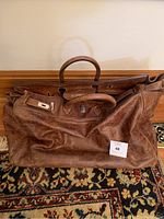 Front view of large tan leather duffel bag on patterned rug showing shape, handles, and padlock hardware