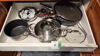 Overview of drawer with assorted cookware including pots, pans, lids, a steamer insert, and a multi-section ceramic serving dish.