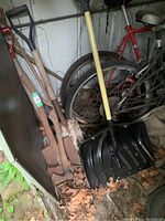 Photo showing two axes with wooden handles leaning against a wall among other stored items with a yellow-handle snow shovel in front.