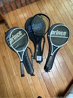 Photo of three tennis rackets, two Prince and one Dunlop, each with its own soft protective case displayed on a wooden floor.