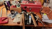 Photo showing a variety of hand tools including hammers, locking pliers, toolboxes, tape rolls, and safety goggles on a wooden table.