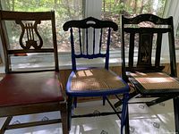 Front view of three antique wooden chairs, one with leather seat, one painted blue with cane seat, one black folding chair with cane seat and decoration.