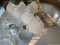 Arrangement of 8 white collapsible paper Chinese lanterns with hooks, 2 champagne glasses, 1 shot glass, Corelli syrup pitcher, metal cooling racks, white ceramic dishes, and a white ladle on a table.