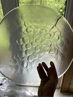 Glass serving plate with textured grape cluster motif held up to window to show translucent quality and grape design