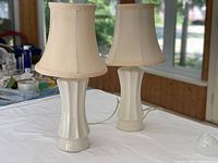 Front view of both white ceramic bedside lamps with fabric bell-shaped shades on a white table.