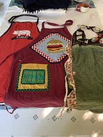Three aprons laid out on table: red apron with Hamlet theme, burgundy apron with hamburger patch and 'Don't Bug Me' green patch, green apron with ruffle edges.