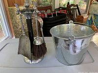 Photo of stainless steel fireplace tool set with four tools on a decorative stand and a galvanized metal bucket with wooden handle next to it.