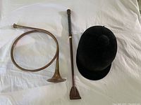 View of antique French horn, vintage black velvet riding helmet, and leather crop laid on white fabric for lot overview.