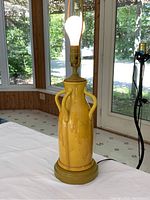 Vintage mustard ceramic mid century modern table lamp with two handles, brass socket, on wooden base, set in front of window with lush greenery visible outside.