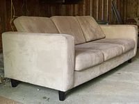 Front-side angle view showing the full length of the beige microfibre couch with three back cushions and three seat cushions, with some visible stains and short black legs.