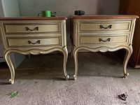 Pair of antique Drexel night tables showing front view with wear on cream paint and wood tops, two drawers each with brass handles