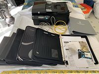 Photo showing the HP Officejet 4500 printer, four black binders, an HP 901 ink cartridge box, thumb tacks, paperwork and cables on a table.