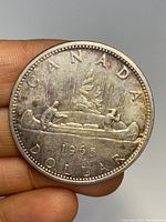 Close-up image of the 1965 Canadian silver dollar coin held between fingers, showing the front design with two figures paddling a canoe and the inscription 'CANADA DOLLAR 1965'.