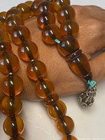 Close-up showing amber beads with a tapered terminating bead and silver-tone charm attached to a teal string.