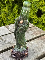 Front view of the dark green carved jade figure of a standing woman on an intricately carved dark wood stand with small coral flower decorations.