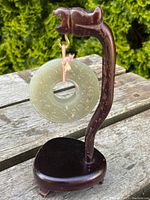 Antique translucent light green jade disc suspended from a curved carved wooden stand, shown on an outdoor wooden surface.