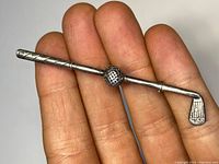 Close-up front view of the silver golf club pin held in hand, showing golf ball detail and overall design.