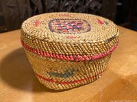 Left side angle of a medium-sized hand-woven basket with lid showing natural and colored fibers and painted patterns.