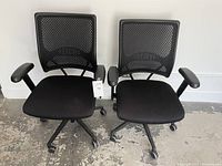 Photo of two black office desk chairs with mesh backs side by side on a concrete floor.