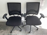 Two black office desk chairs side by side on concrete floor against white wall, showing mesh backs, cushioned seats, adjustable armrests, and caster wheels.