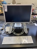 Front view of Apple Cinema HD Display monitor with cables, Apple wired keyboard, and a clear acrylic laptop stand on a black surface.