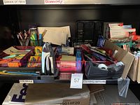 View of a black shelf with assorted office supplies including markers, pens, pencils, crayons, tape dispensers, binder clips, papers, and a calculator in a cardboard sorter box.