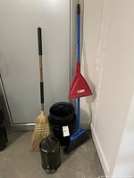 Photo shows four tall black plastic garbage bins stacked inside each other, one small black metal garbage bin with lid placed in front, a natural fiber broom and a blue plastic broom with attached dustpan leaning against a white cupboard and wall.