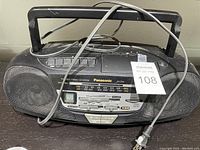 Front view of Panasonic boombox with speaker grills, control buttons, radio tuner, cassette player door, handle, power cord and paint splatters.