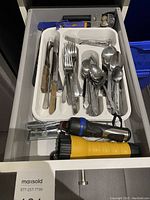 Drawer with assorted cutlery, two flashlights, can opener, and other metal utensils in a white tray.