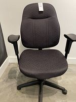 Front view of black fabric padded office chair with adjustable armrests and five black caster wheels on a concrete floor.