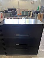 Front view of a black metal horizontal filing cabinet with two drawers in a warehouse setting. Provides clear condition and design details.