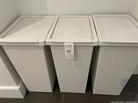 Three white IKEA FILUR storage bins with lids closed, standing side by side on floor against wall.