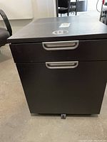 Front view of dark brown/black finishing drawer unit with two silver handles, on wheels, standing on floor.