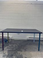 Full view of the Ikea table with dark brown LINNMON tabletop and four metal legs each painted a different color: purple, green, blue, and gray. Table is set against a white wall and floor with visible cords beneath.